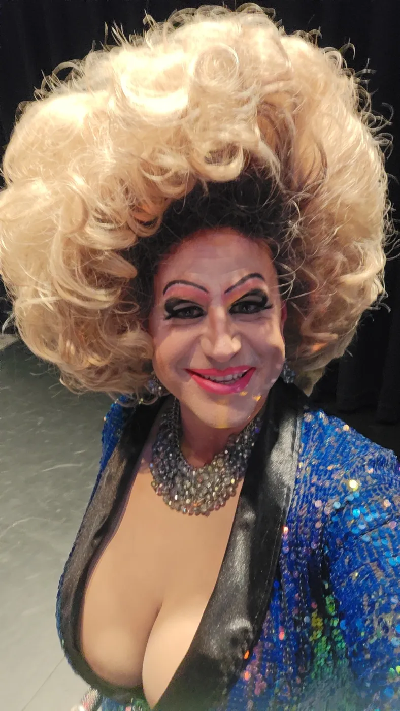 Divinity Masters in a big blonde wig and sparkling blue sequin jacket, smiling